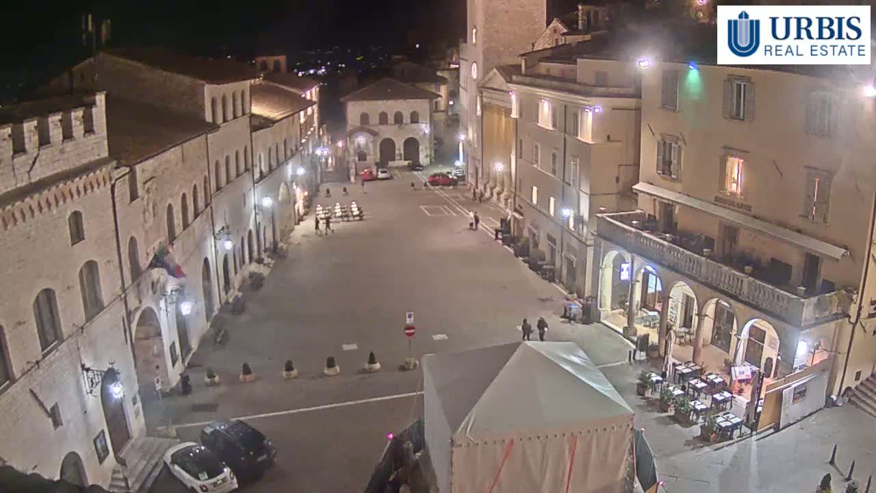 A nighttime, high-angle view of a town square with several buildings, a few cars, and some outdoor seating at a restaurant.