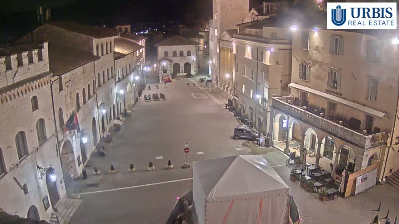 A nighttime, high-angle view of a large, mostly empty town square surrounded by illuminated stone buildings.