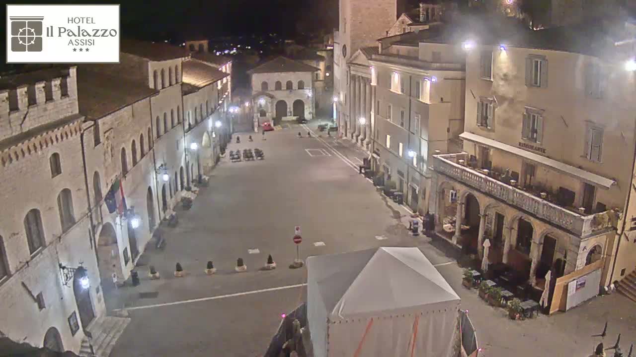 A nighttime, high-angle view of a mostly empty town square surrounded by stone buildings, with a white tent in the foreground.