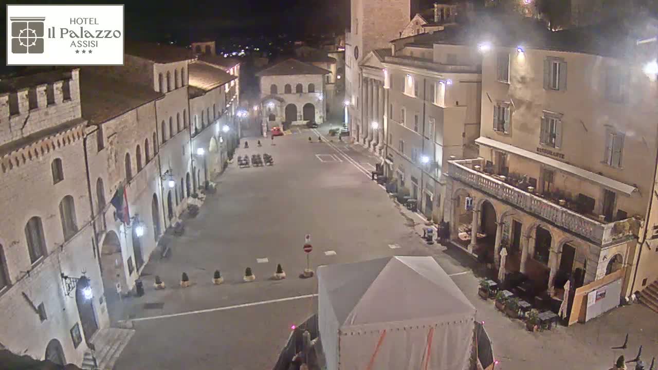 A mostly empty town square at night, illuminated by streetlights, is surrounded by historic-looking buildings.