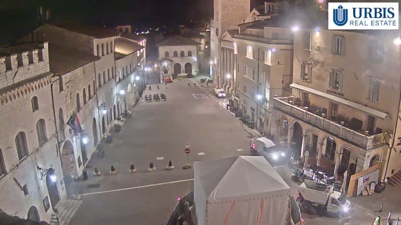 A mostly empty town square at night, surrounded by historic buildings, is illuminated by streetlights.