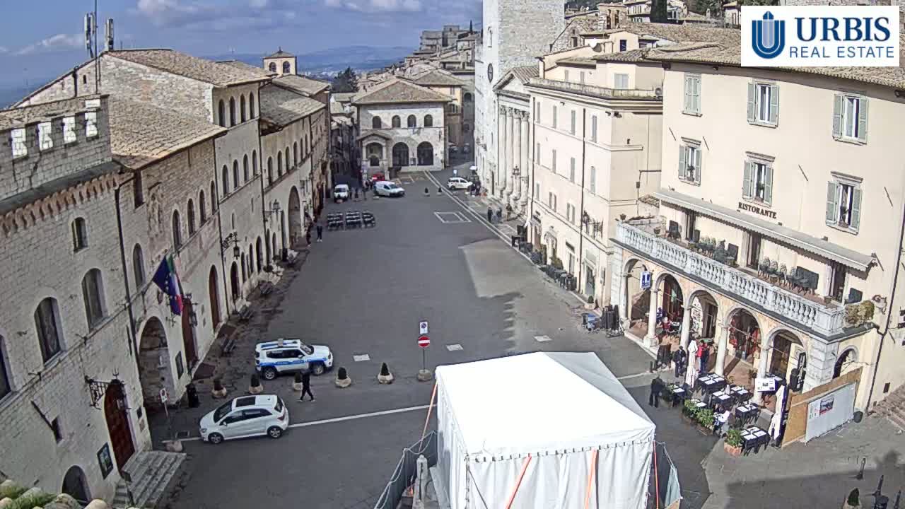 A mostly empty town square in bright sunlight is surrounded by historic-looking buildings, with a few cars and pedestrians visible.