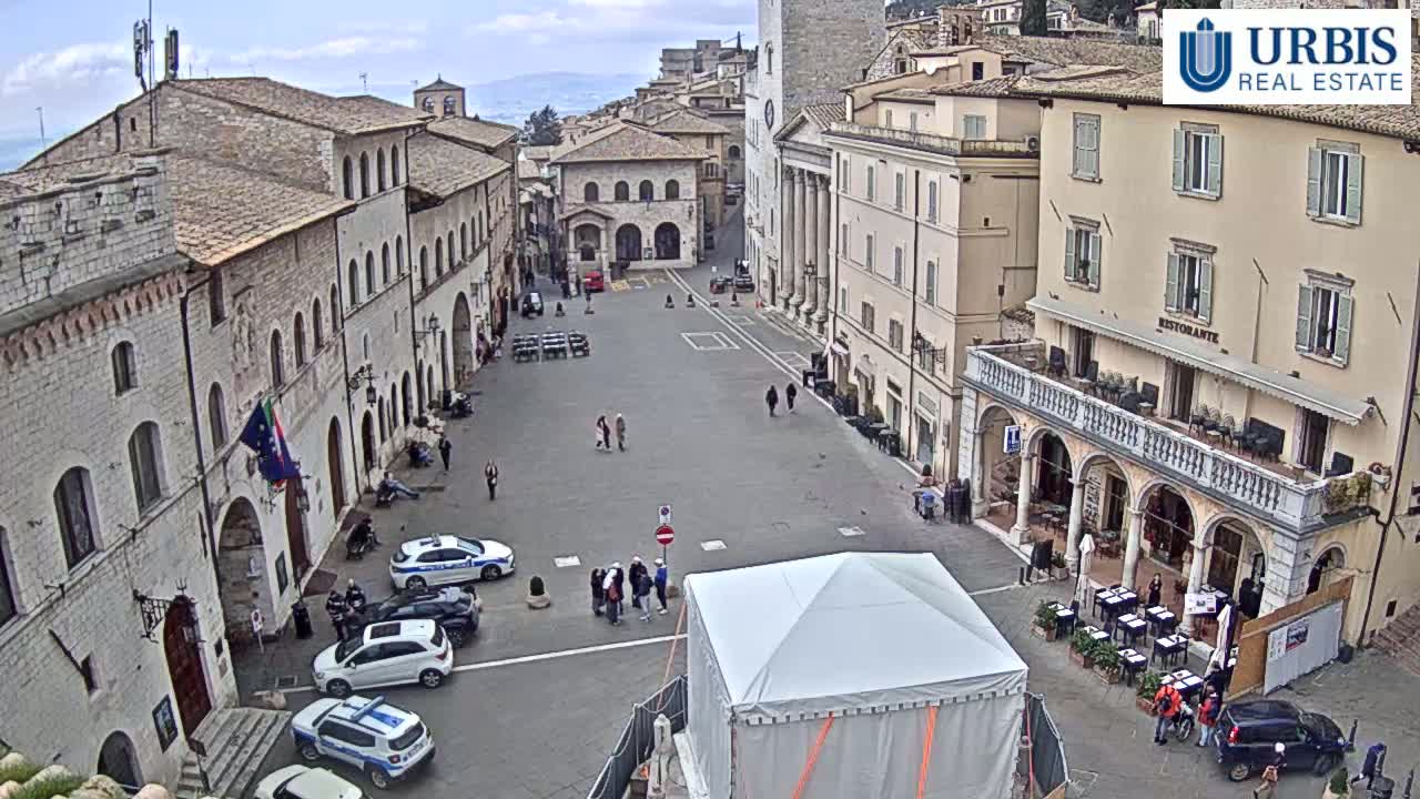 A large, mostly empty town square is surrounded by stone buildings under a cloudy sky, with several people, cars, and a large white tent visible.