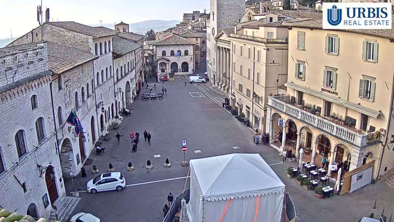 A mostly empty town square with stone buildings, a few pedestrians, and a white tent in the center, under a mostly clear sky.