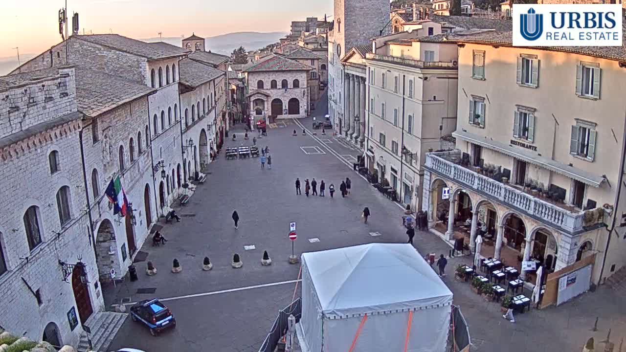 A mostly empty town square with stone buildings, a few people, and a white tent under a clear, sunrise sky.