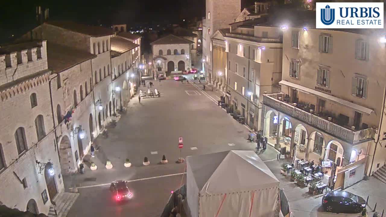 A nighttime, high-angle view of a mostly empty town square surrounded by historic-looking buildings, with a few cars and pedestrians present.