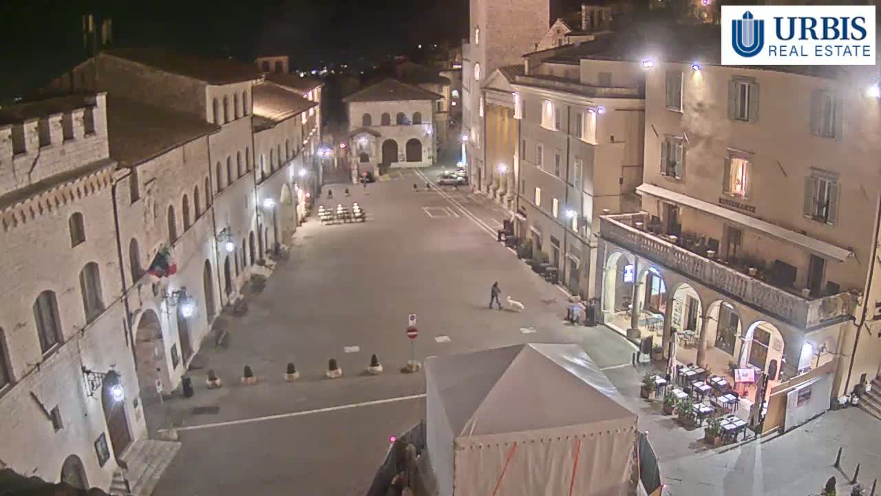 A nighttime, high-angle view of a mostly empty town square surrounded by old, stone buildings, with a few people and a dog visible.