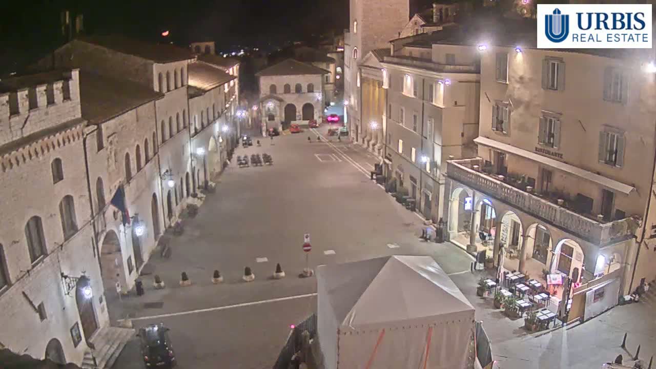 An empty, dimly lit town square at night is surrounded by aged stone buildings, one of which has outdoor seating.