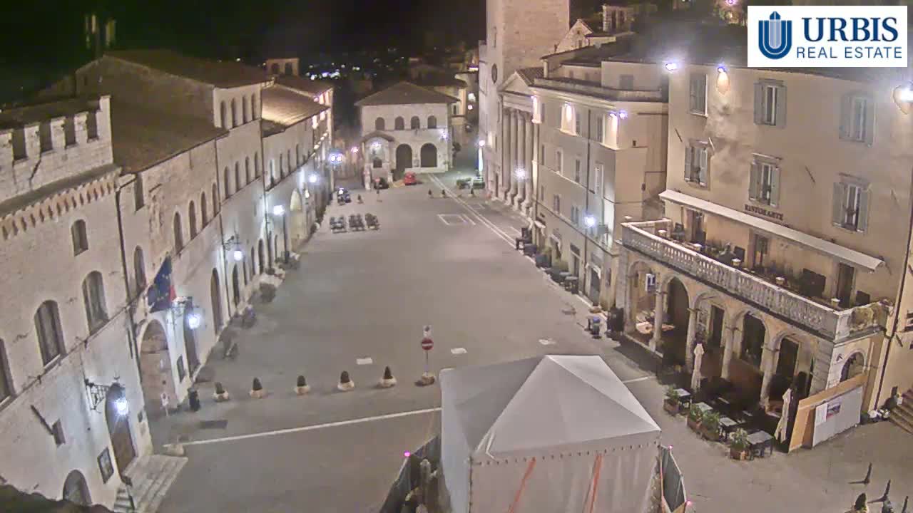 A mostly empty town square at night, illuminated by streetlights, is surrounded by historic-looking buildings.