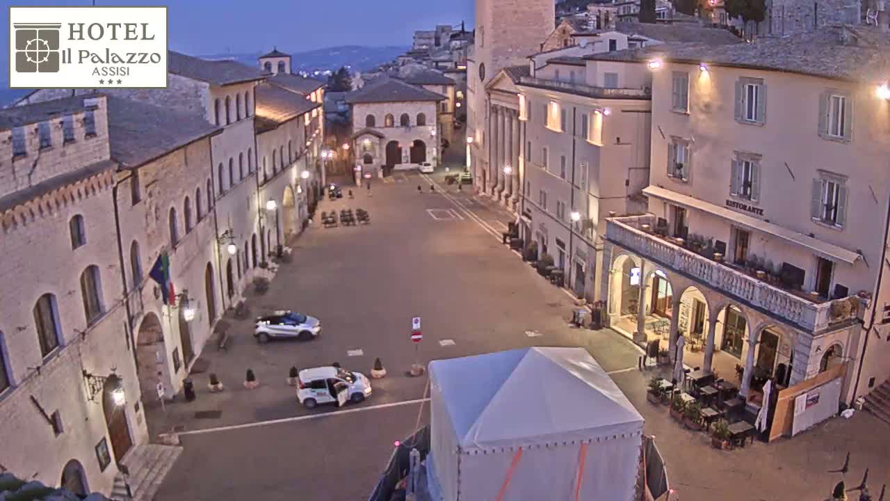 A mostly empty town square at dusk is surrounded by historic stone buildings, with a few cars and a temporary tent visible.