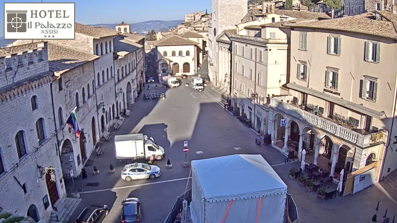 A sunny day in a large town square in Italy, surrounded by historic buildings, with several cars and a covered structure in the center.