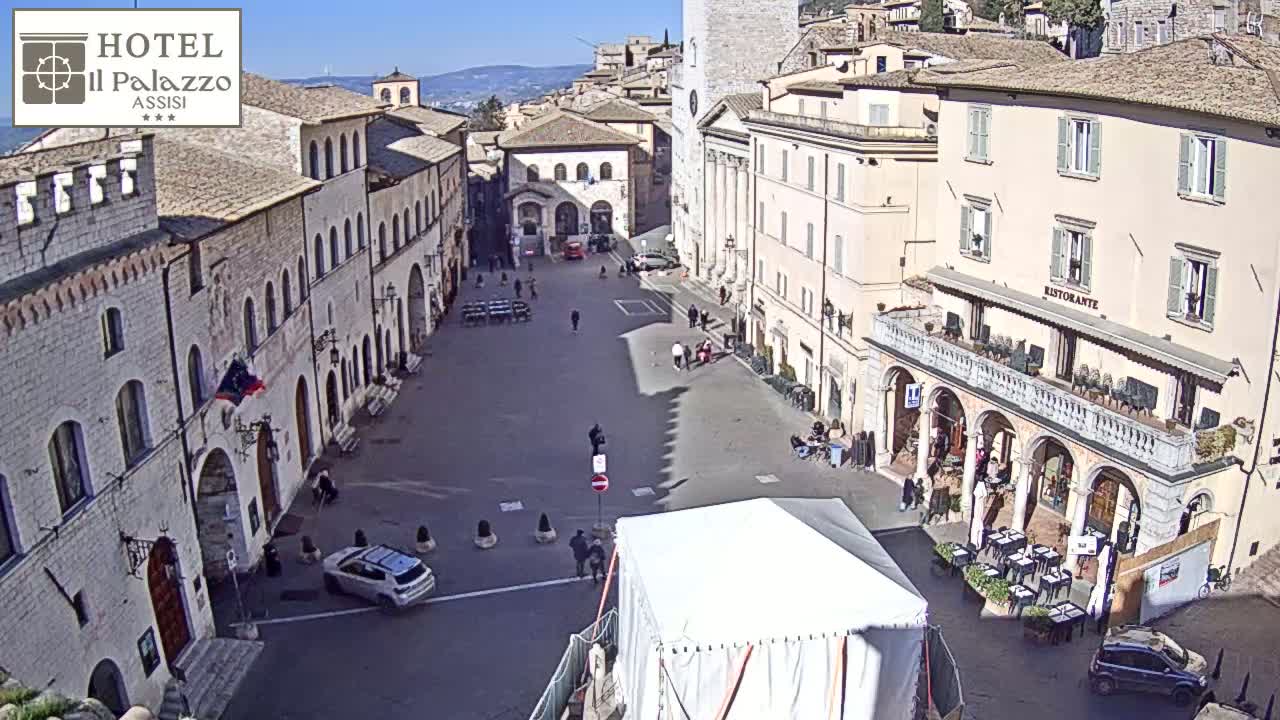 A mostly empty town square in bright sunlight is surrounded by historic stone buildings, with a few people and cars visible, and a white tent set up in the center.