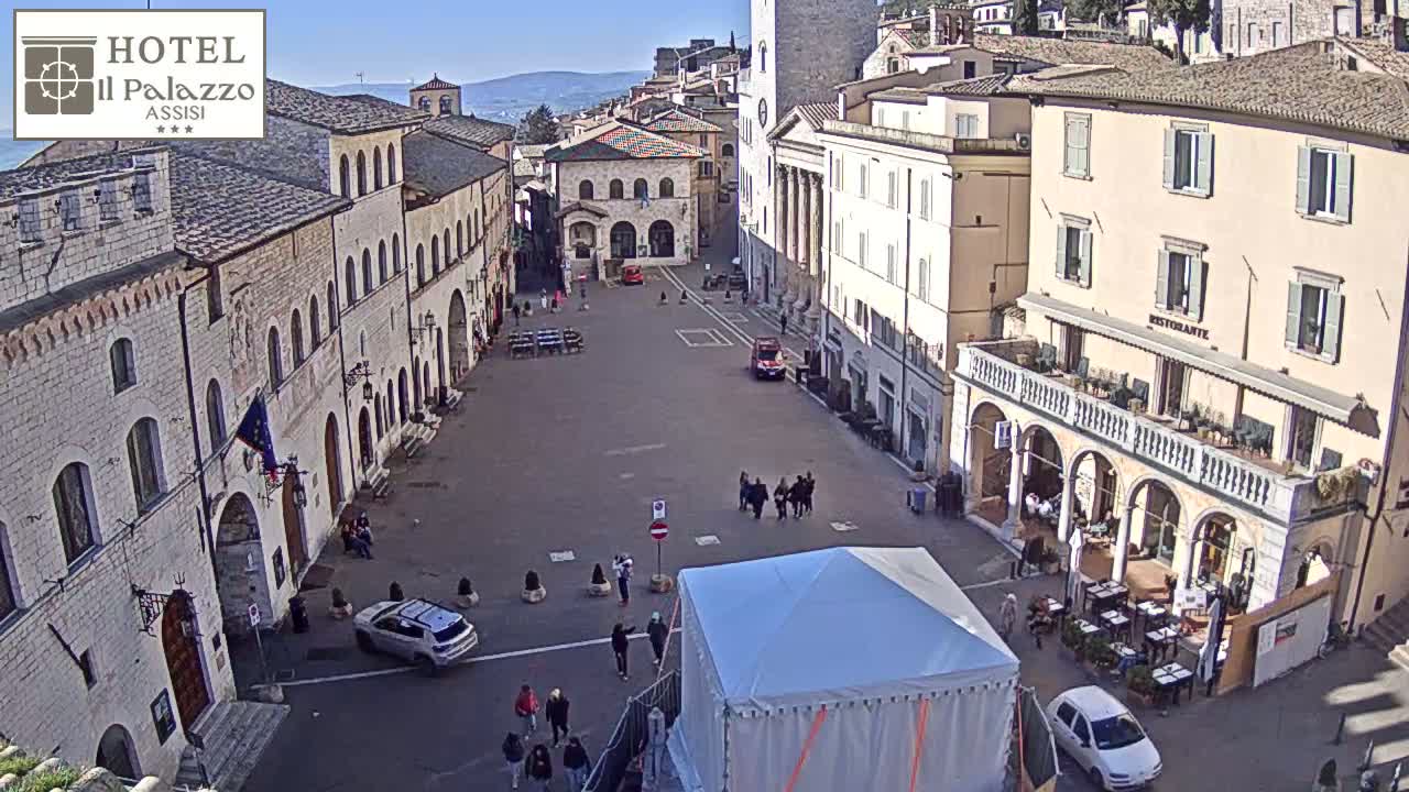 A mostly empty town square in a European city on a sunny day, with several people scattered about and cars parked along the sides.