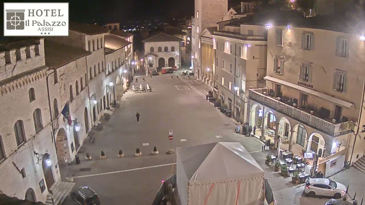A mostly empty town square at night, illuminated by streetlights, is surrounded by historic-looking buildings.