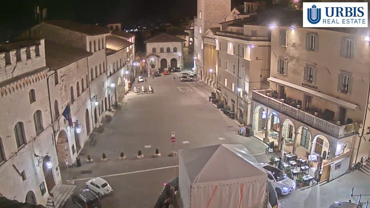 A mostly empty town square at night, surrounded by historic-looking buildings, is lit by streetlights.