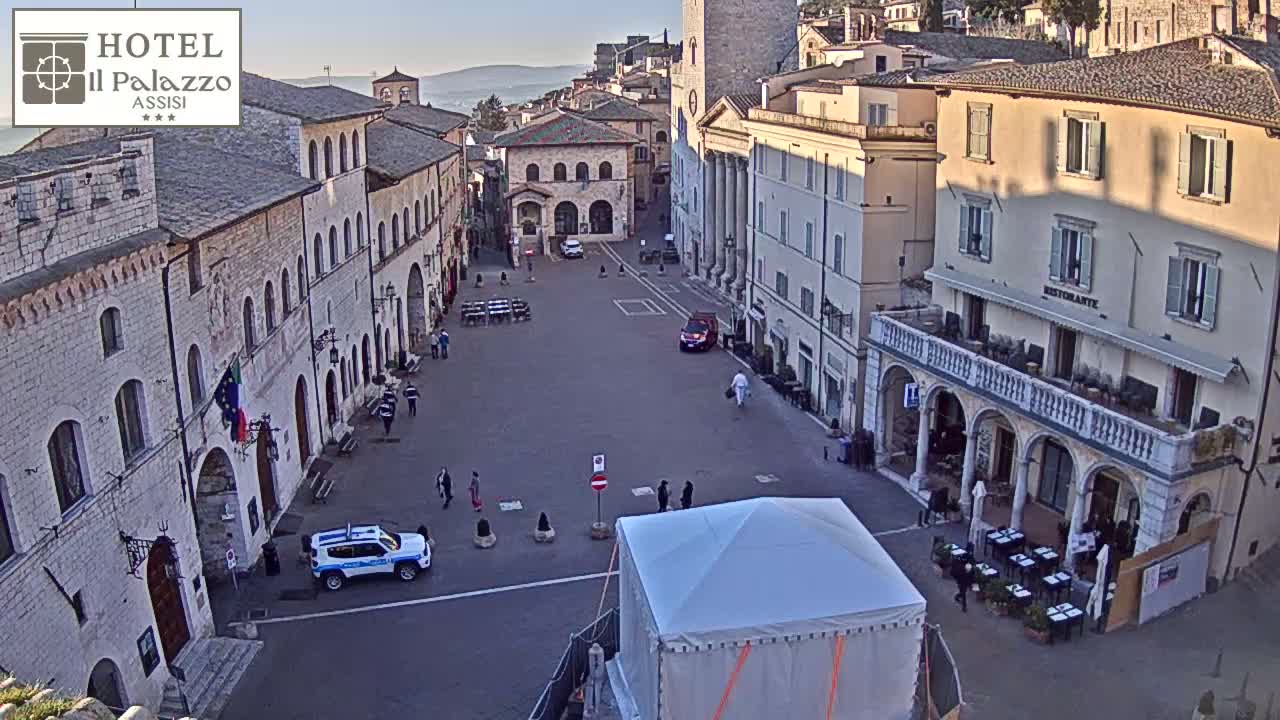 A mostly empty town square, under a clear sky, is surrounded by aged stone buildings, with a few people, some vehicles, and outdoor seating.