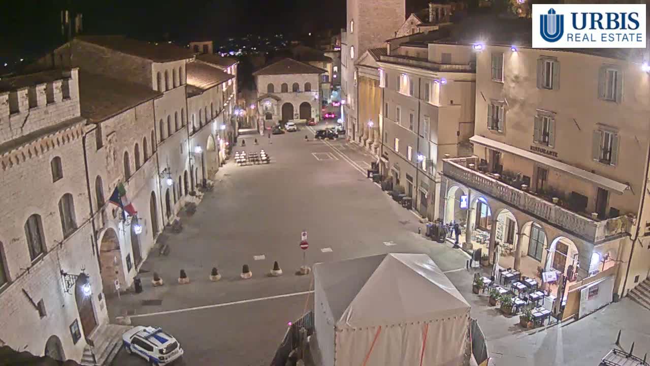 An empty town square at night, surrounded by stone buildings, is illuminated by streetlights.