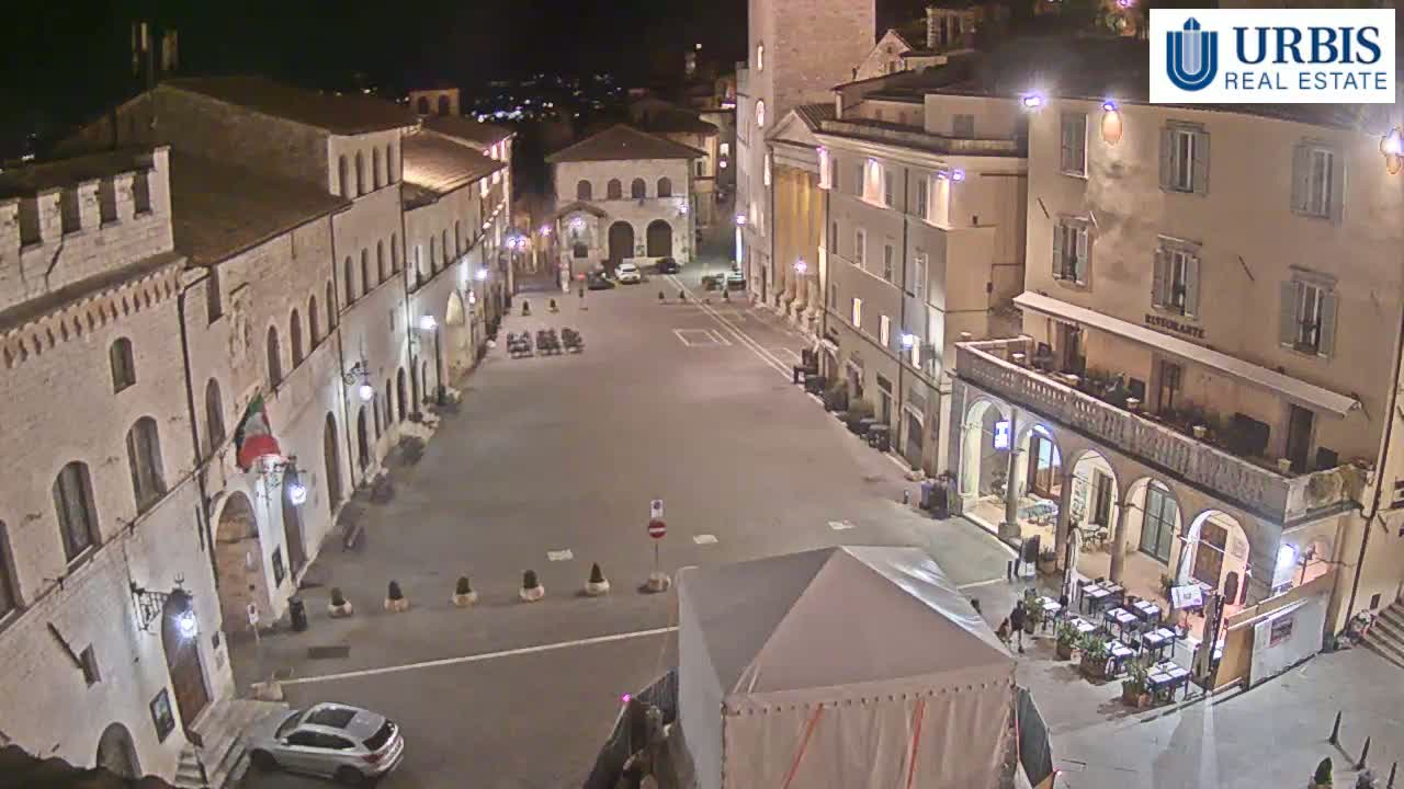 A mostly empty town square at night, surrounded by buildings, with a few cars and outdoor seating at a restaurant.