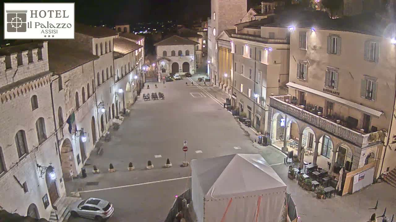 A mostly empty, stone plaza at night, bordered by buildings, with a few cars and a small tent visible.
