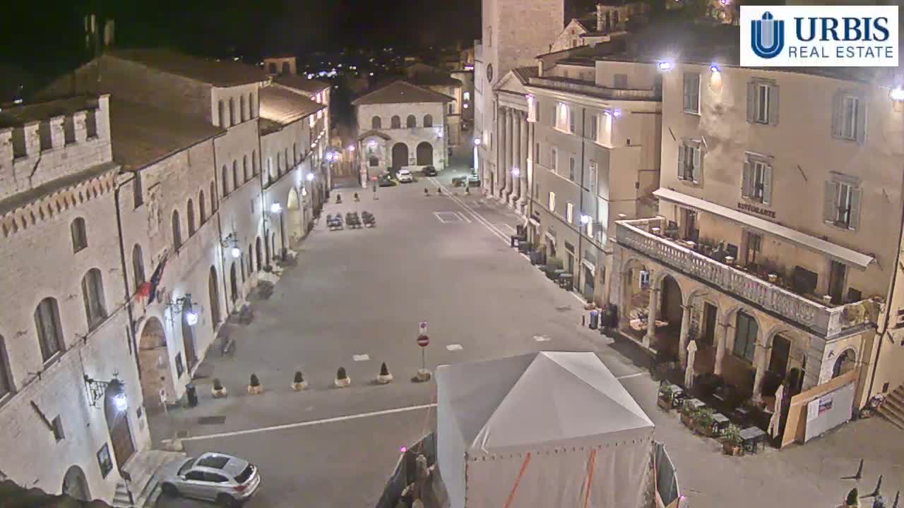 A mostly empty town square at night, surrounded by old stone buildings, is illuminated by streetlights.