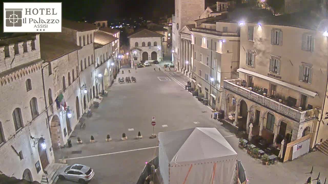 A mostly empty town square at night, surrounded by historic-looking buildings, is illuminated by streetlights.