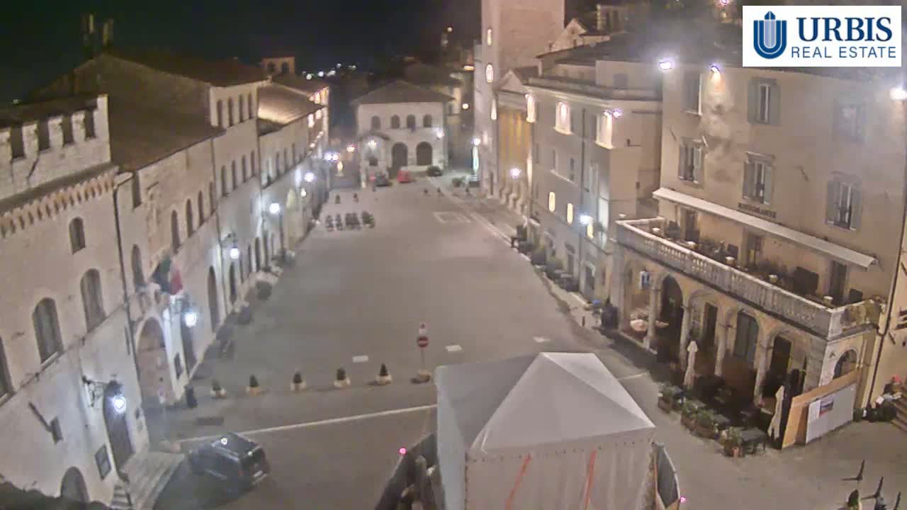 A mostly empty town square at night, surrounded by historic-looking buildings, is illuminated by streetlights.