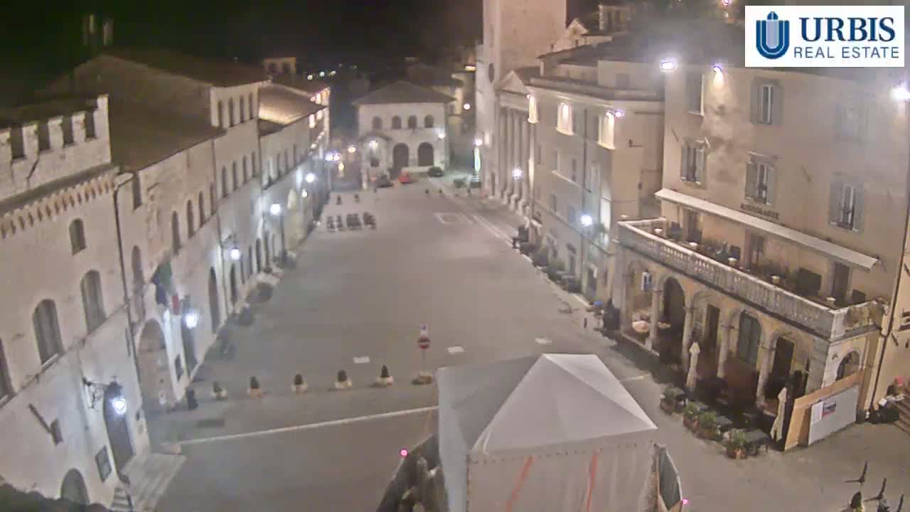 A mostly empty town square at night, surrounded by historic-looking buildings, is illuminated by streetlights.