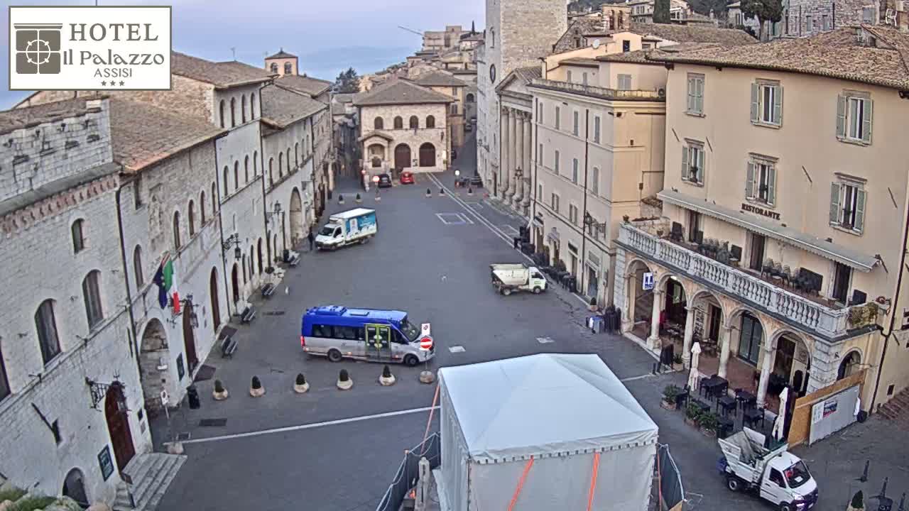 A mostly empty town square in a European-style town, under an overcast sky, features several parked vehicles and buildings along its sides, with one featuring outdoor seating.