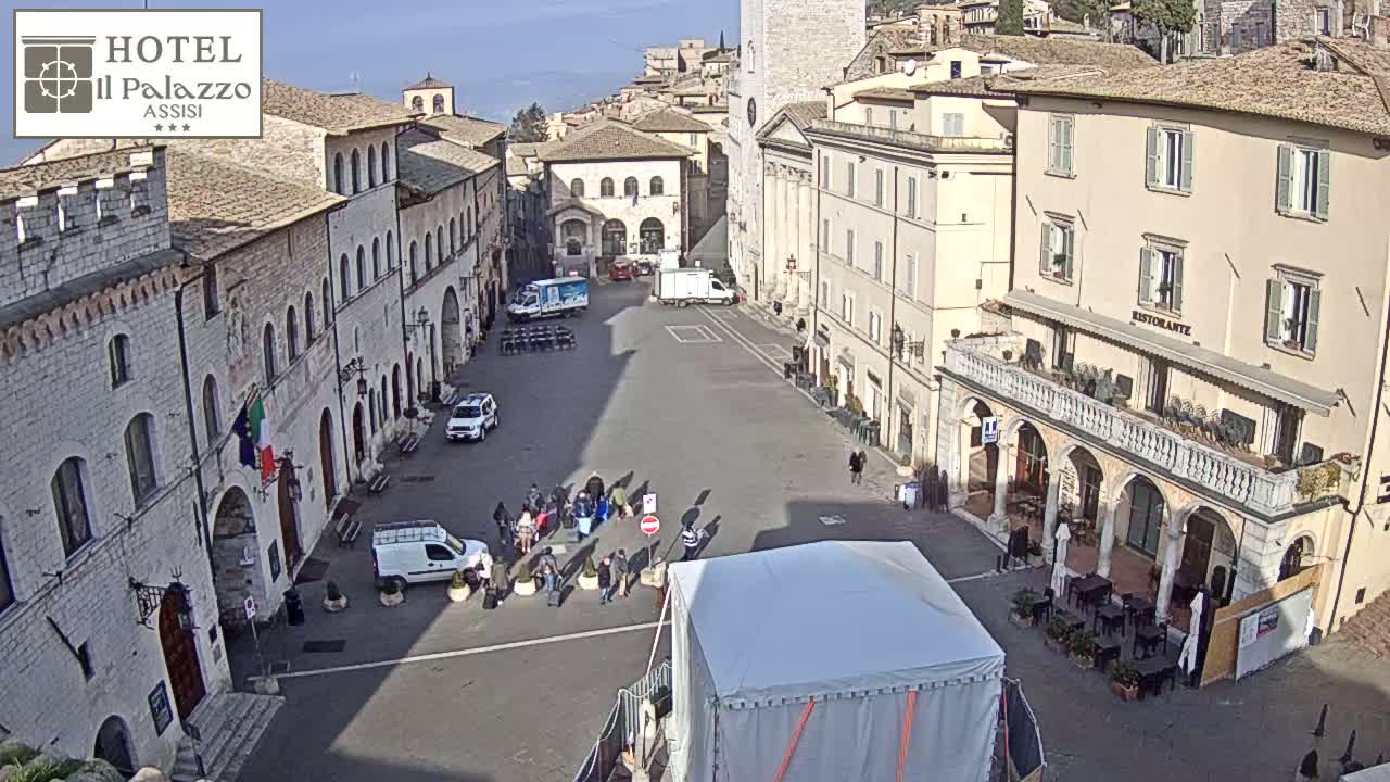 A sunny day in a large, paved town square surrounded by aged, light-colored buildings, with several people and a few vehicles present.