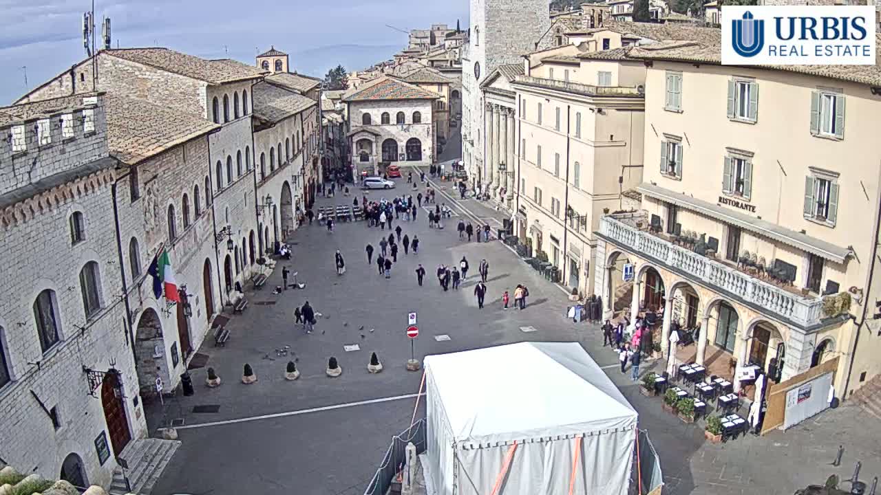 A large town square with several stone buildings, a few people walking around, and a white tent in the center, under a clear, bright sky.