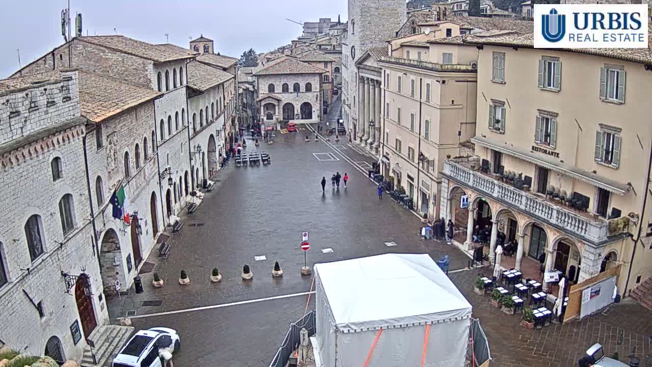 A wet, empty town square is surrounded by stone buildings under an overcast sky.