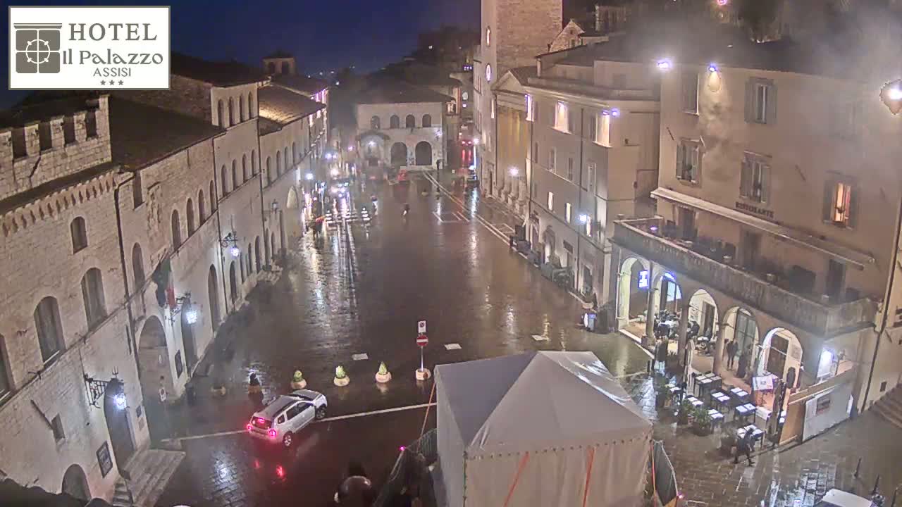 A rain-slicked town square at night, surrounded by historic buildings, with a few cars and people sparsely scattered about.