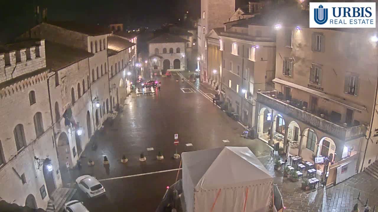 A wet, empty town square at night is surrounded by stone buildings, with a few cars parked and a white tent in the center.