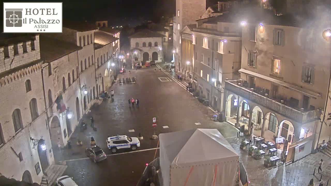 A wet, empty town square at night is surrounded by old, stone buildings with lights illuminating the scene.