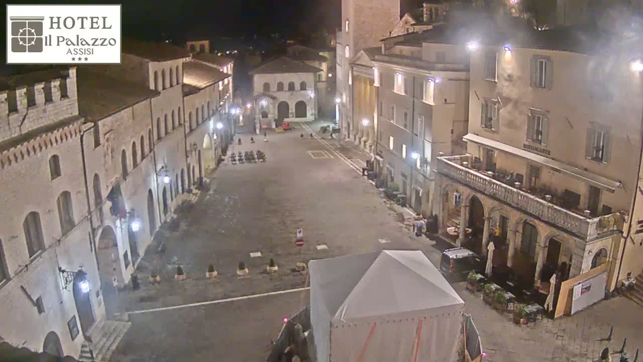 An empty, stone plaza at night, surrounded by old buildings, is dimly lit under a hazy sky.