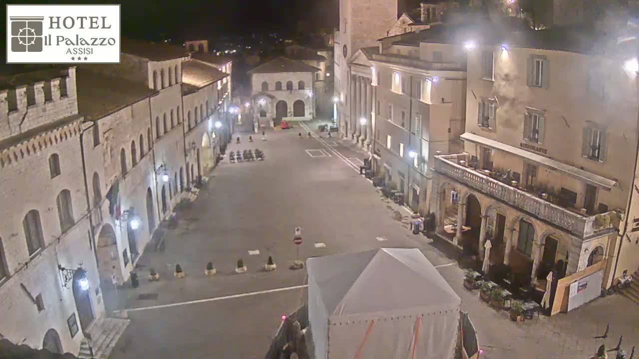 A mostly empty, paved town square at night, surrounded by stone buildings, with a large white tent in the foreground.