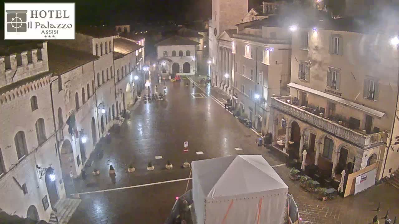 A rain-slicked, empty town square at night is surrounded by old, stone buildings lit by streetlights.