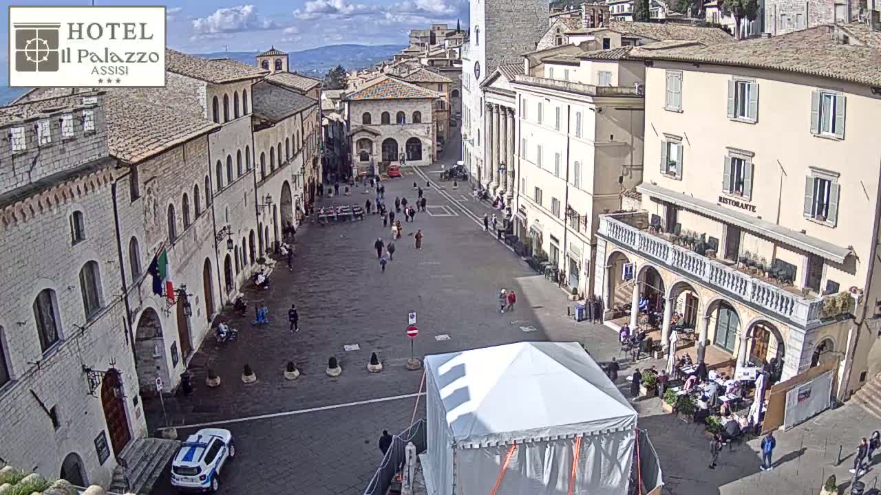 A mostly sunny, outdoor view of a large town square in Italy, with several people walking around, some buildings with arched entries, and a white tent in the center.