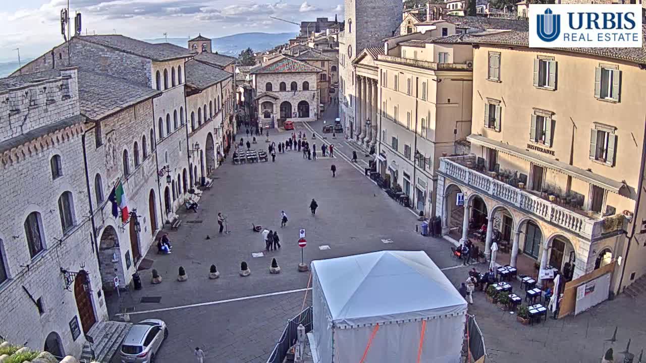 A mostly empty town square in a European town, under a partly cloudy sky, is surrounded by stone buildings, with a few people scattered about and a large white tent in the center.