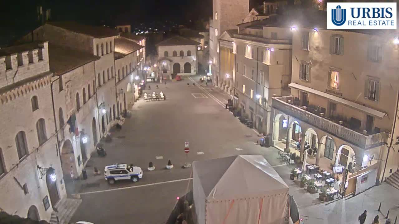 A nighttime, high-angle view of a mostly empty town square surrounded by stone buildings, with a single police car parked and a white tent set up in the center.