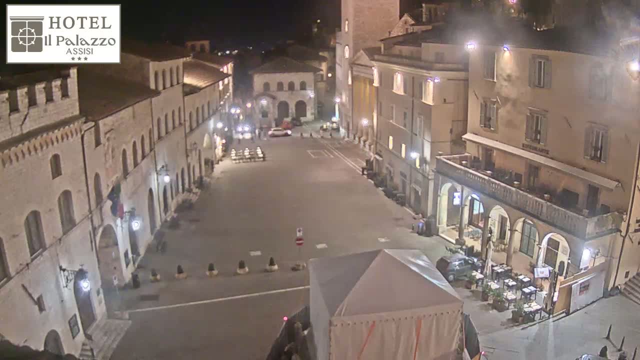A nighttime, high-angle view of a mostly empty town square surrounded by historic stone buildings, with a few cars and outdoor seating at a restaurant.