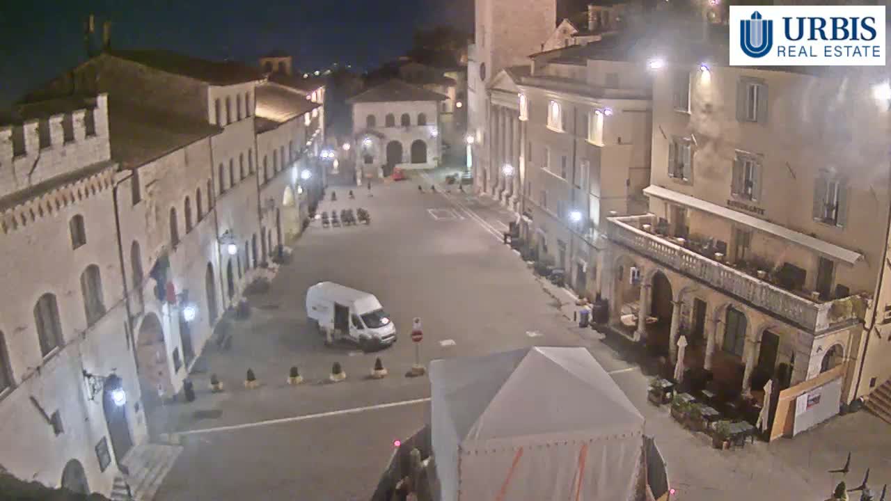 A mostly empty town square at night, surrounded by historic-looking buildings, is illuminated by streetlights.