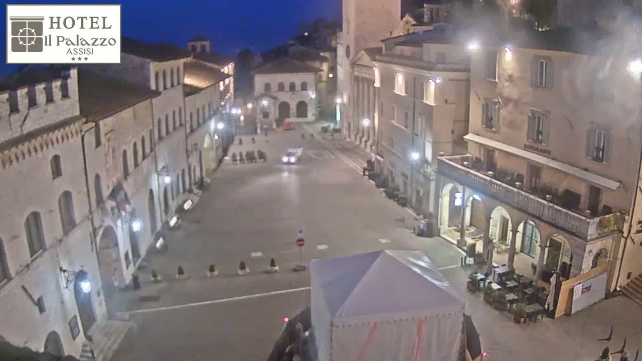 A mostly empty town square at night, lit by streetlights, is surrounded by several-story buildings.