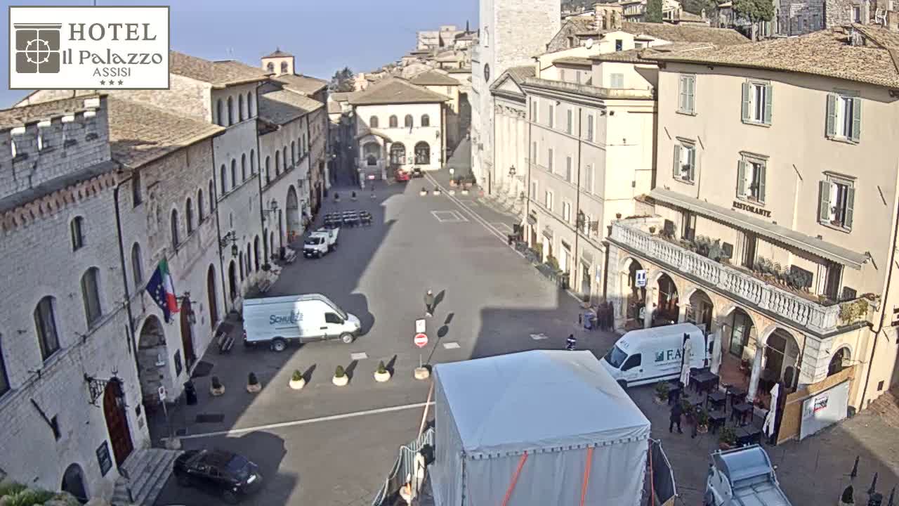 A sunny day reveals a large town square in Italy, surrounded by historic buildings, with several vehicles and a temporary tent structure.