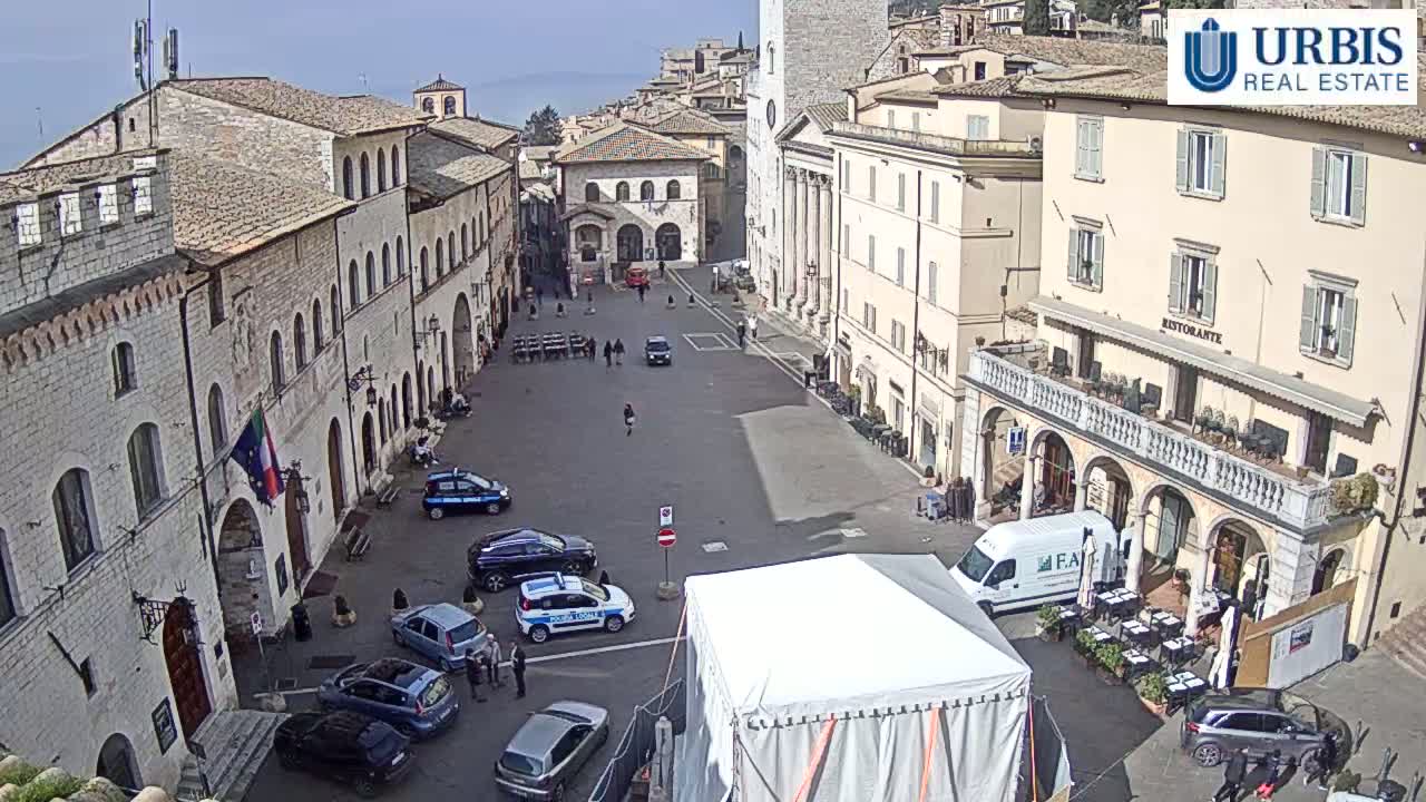 A large town square with several cars, a white tent, and stone buildings surrounds it under a clear, sunny sky.