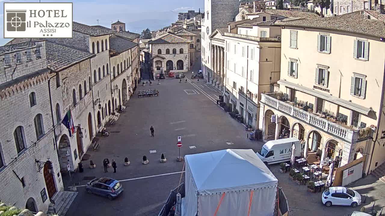 A mostly empty town square in bright sunlight is surrounded by historic stone buildings, with a few people and cars visible.