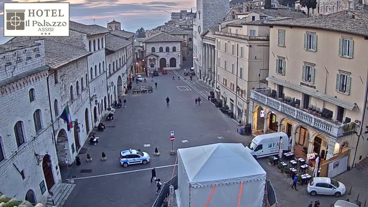 A mostly empty town square, surrounded by aged stone buildings, is viewed from above under a twilight sky.
