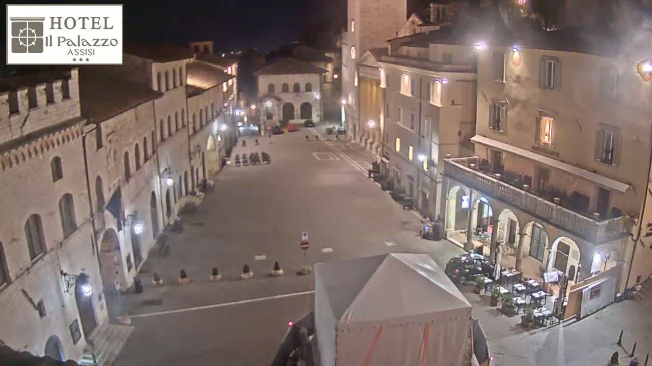 A mostly empty town square at night, illuminated by streetlights, is surrounded by historic-looking buildings.