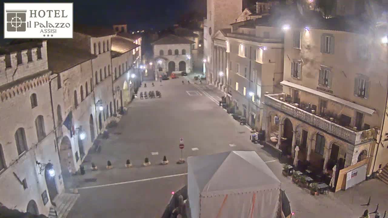 A mostly empty town square at night, lined with stone buildings, is illuminated by streetlights.