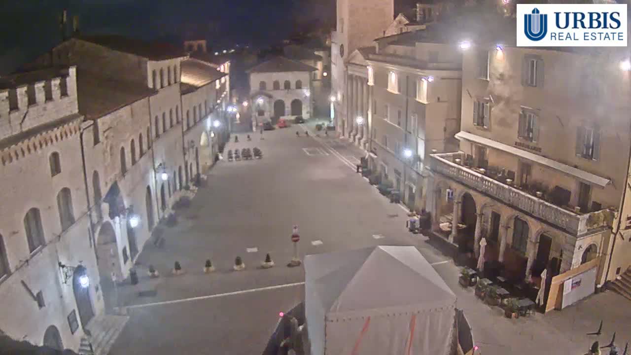 A mostly empty town square at night, surrounded by old stone buildings, is lit by streetlights.